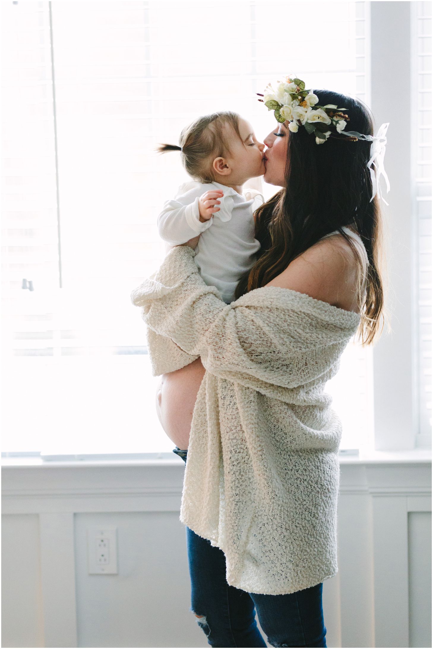 mom in a flower crown and little girl kiss in front of a window | maternity session