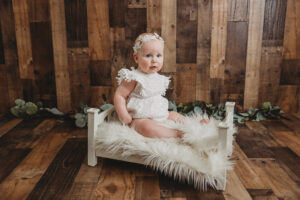 baby dressed in white with a headband sitting on a miniature bed